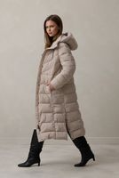 Frau trägt beige, knielanges Puffercoat mit schwarzem Stiefel, lässig stehend.