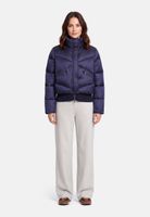 Frau trägt navy-blauen Puffer-Jackenanzug mit hochgeschlagenem Kragen, helle Hosen und flache Schuhe.