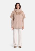 Frau trägt beige, flauschigen Poncho mit hochgeschlagenem Kragen, weiße Hose und flache Schuhe.