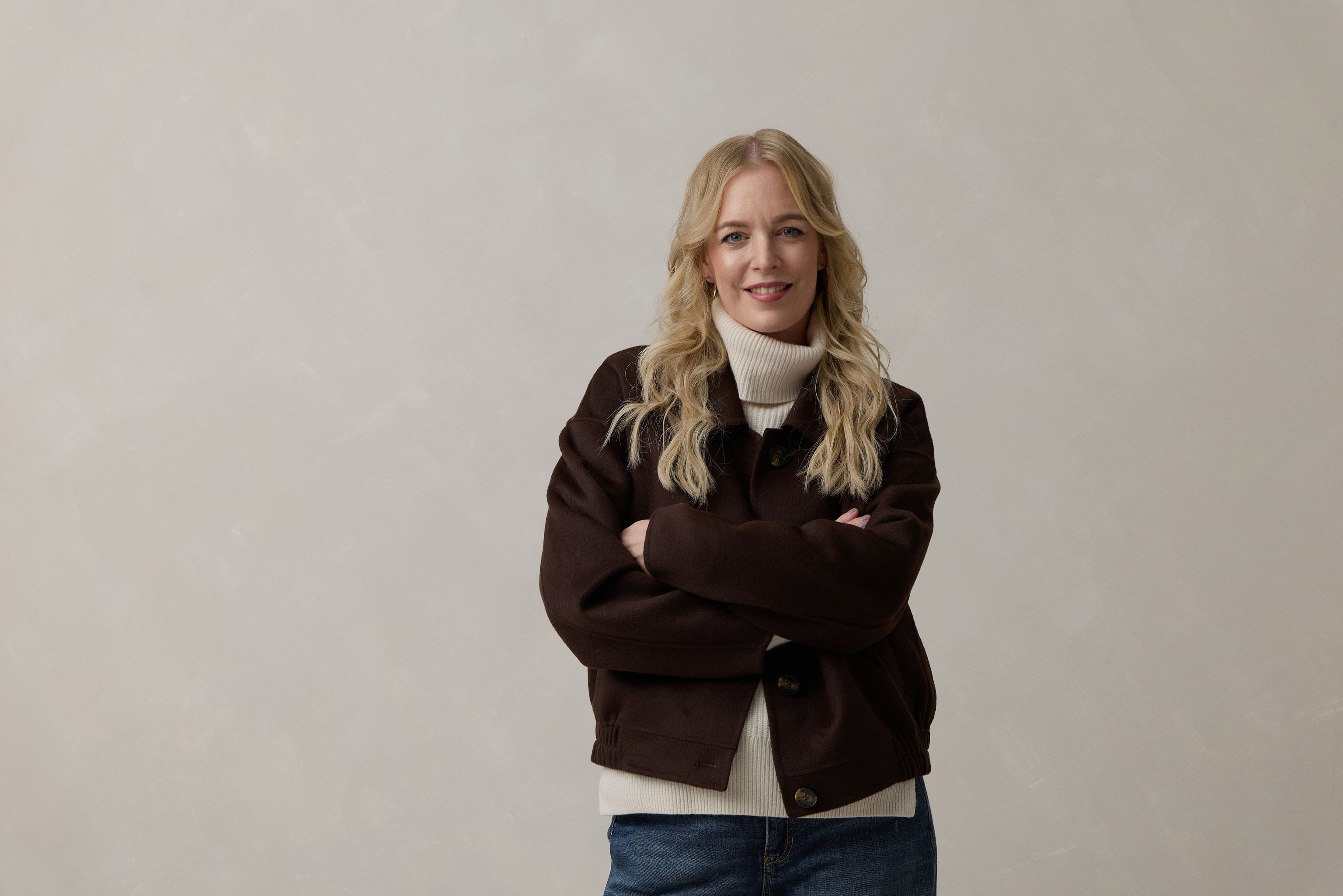 Frau mit blonden Locken trägt einen braunen oversize-Pulli und lächelt selbstbewusst.