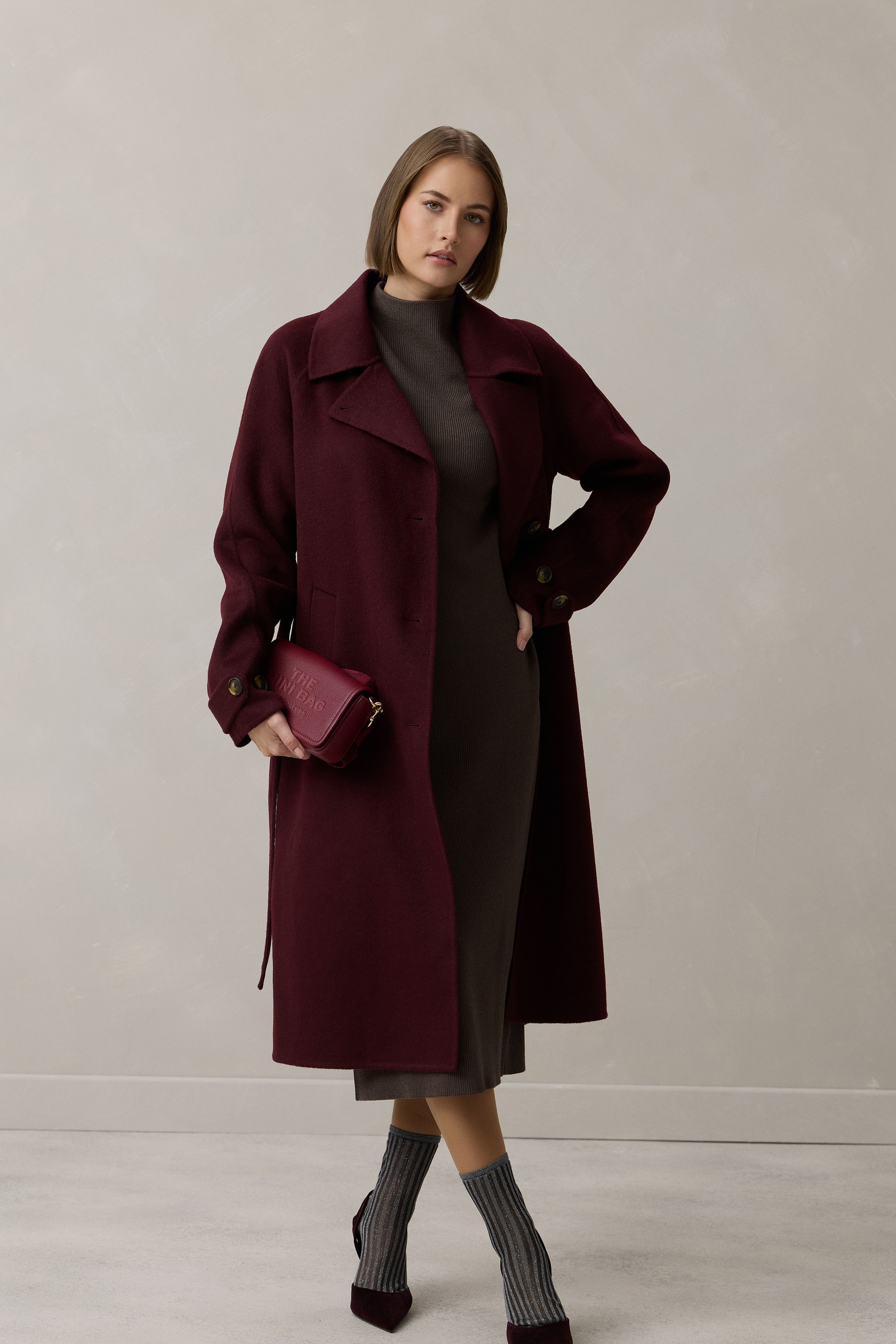 Elegante Frau in burgunderfarbenem Mantel, grauer Strickkleidung und Stiefeln mit Handtasche.
