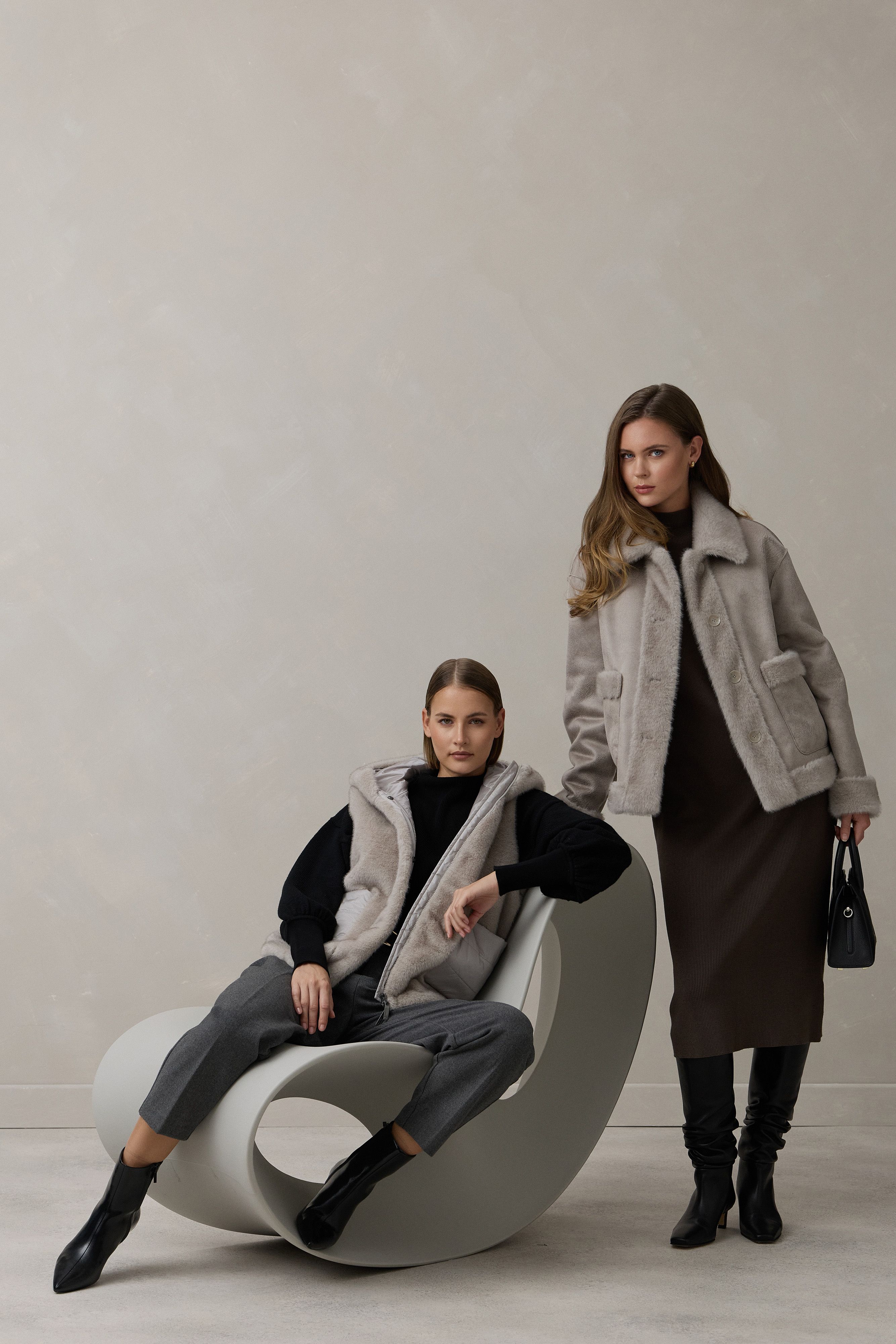 Zwei Frauen in stylischen Winteroutfits auf modernem Möbelstück vor neutralem Hintergrund.
