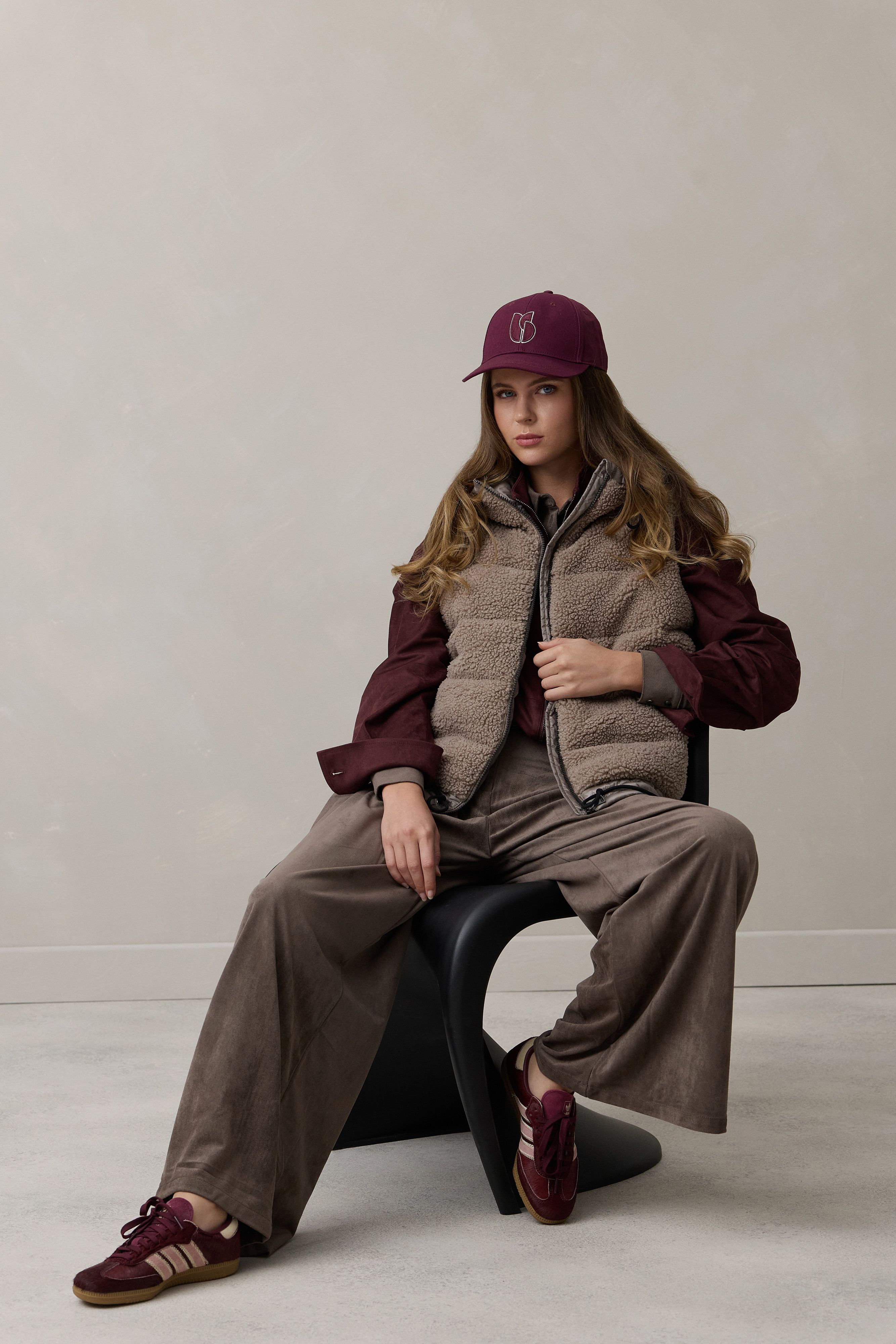 Person sitzt lässig in oversized Jacke, Cap und weiten Hosen, marone Schuhe, neutraler Hintergrund.